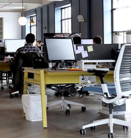 A chair sits empty behind a computer in a room full of desk workers
